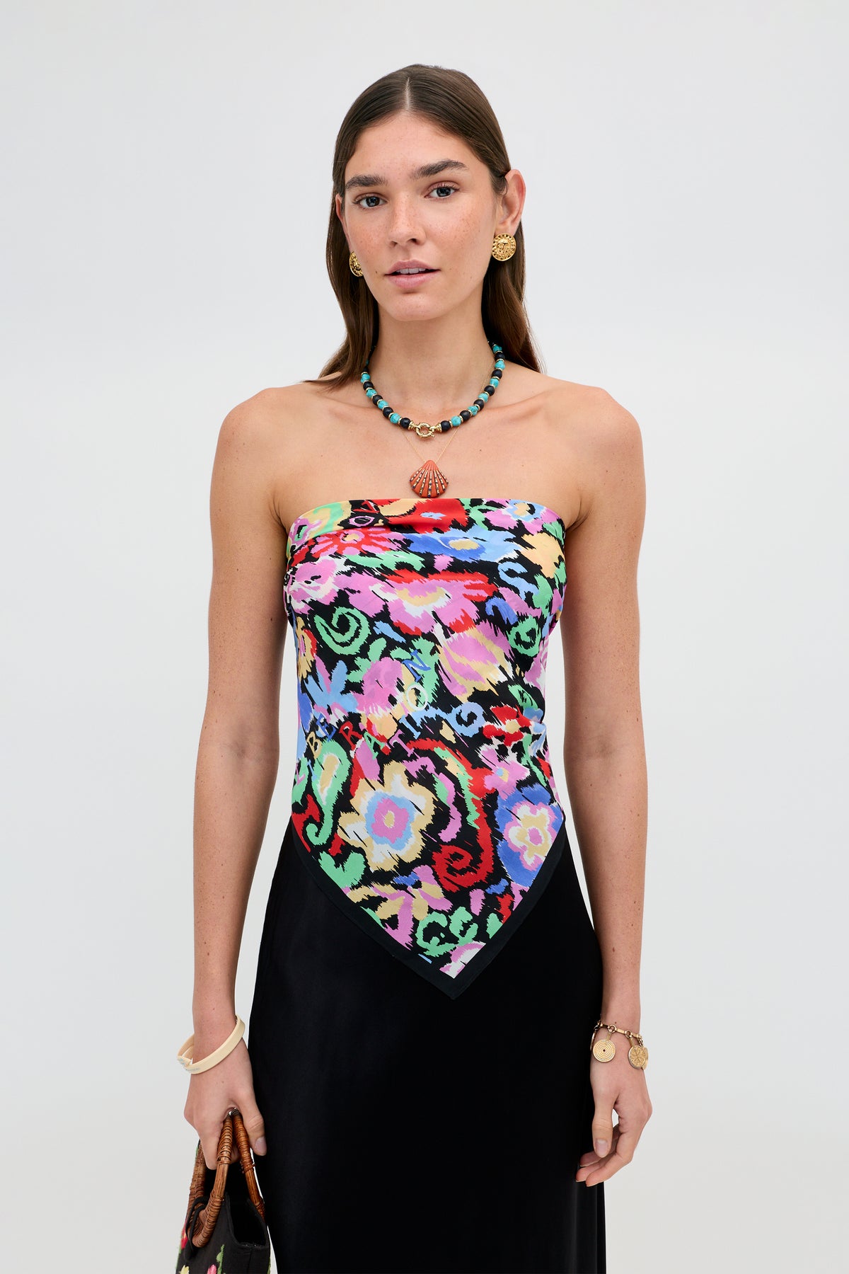 A woman stands against a plain background, wearing the Menno - Ikat Bloom strapless top—a colorful, patterned piece—paired with a black skirt. She has long brown hair, gold earrings, a beaded necklace, and a bracelet, with a neutral expression.