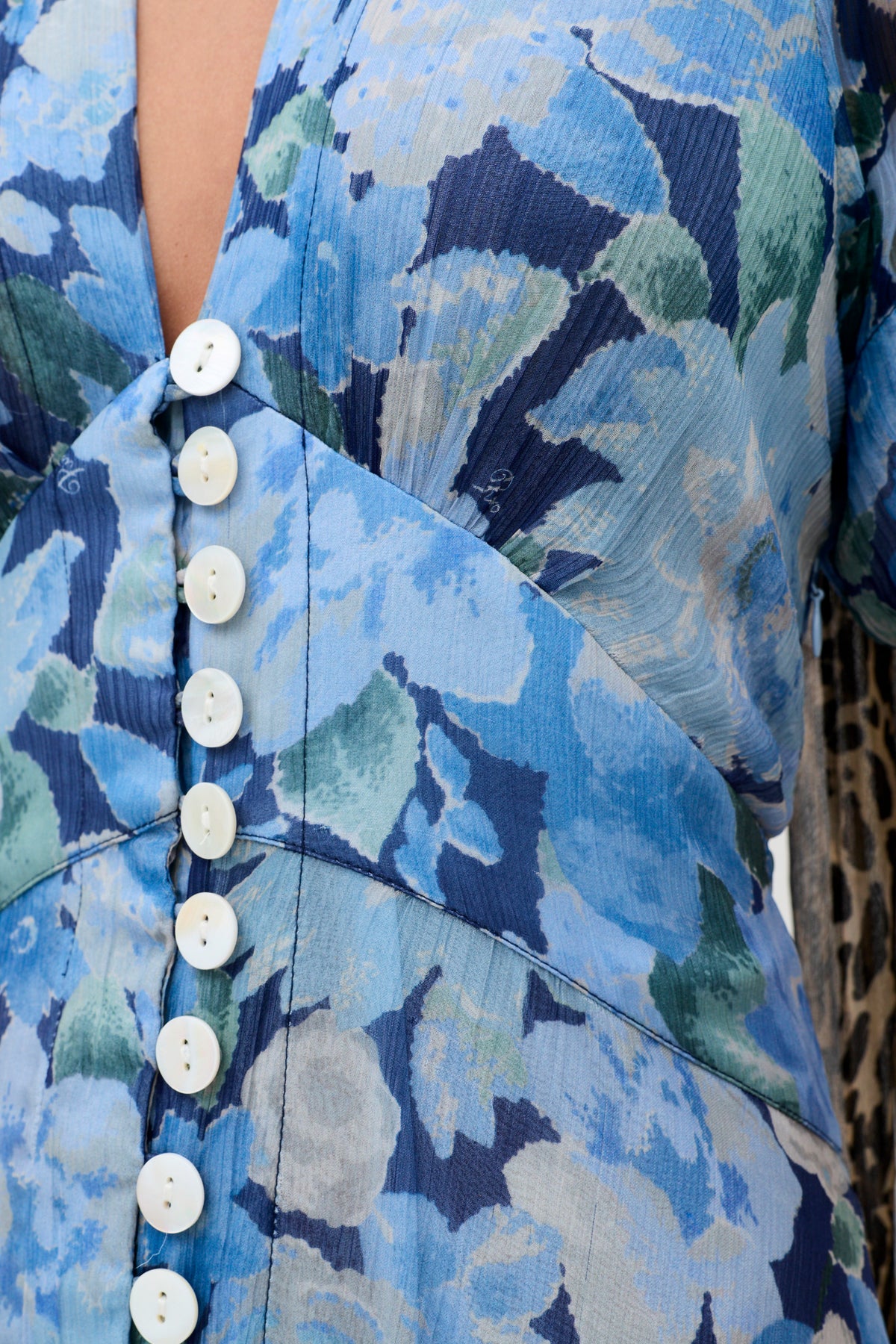Close-up of the novara dress chelsea floral blue, showcasing white buttons along a diagonal seam. The fabric displays various shades of blue and green flowers, with part of an animal print garment visible on the right side.