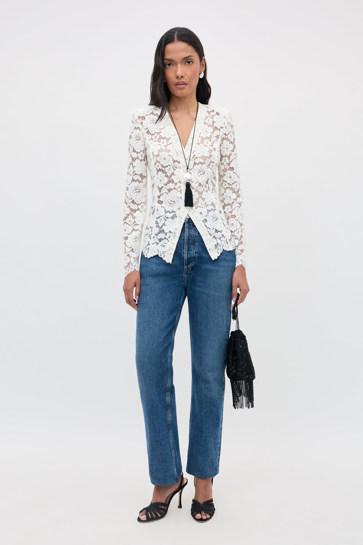 A woman stands against a plain background wearing the Padma - Cream lace blouse, blue jeans, black heeled sandals, and holding a small black fringed purse. She has long dark hair and wears a black tassel necklace.
