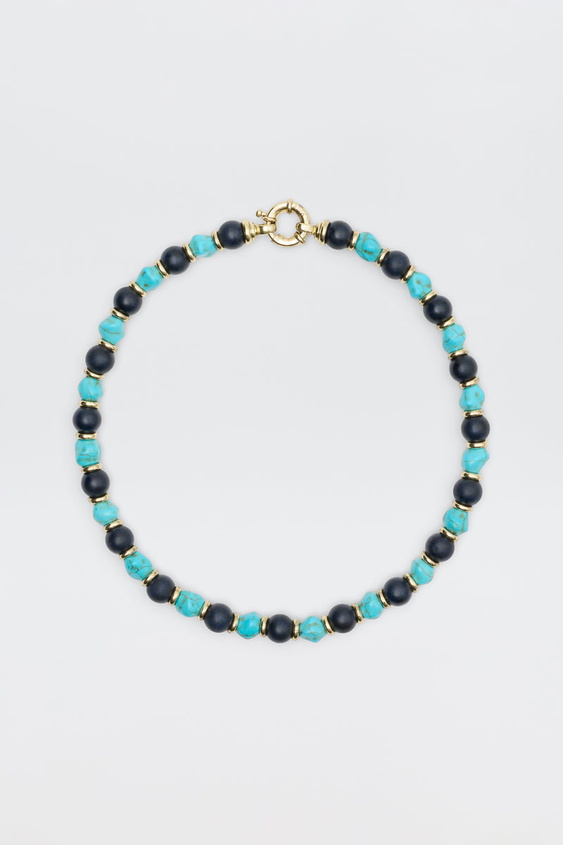 The "Palmara - Blue" necklace features alternating blue-green and dark blue round beads with gold spacers and a gold spring ring clasp, elegantly displayed against a plain light background.