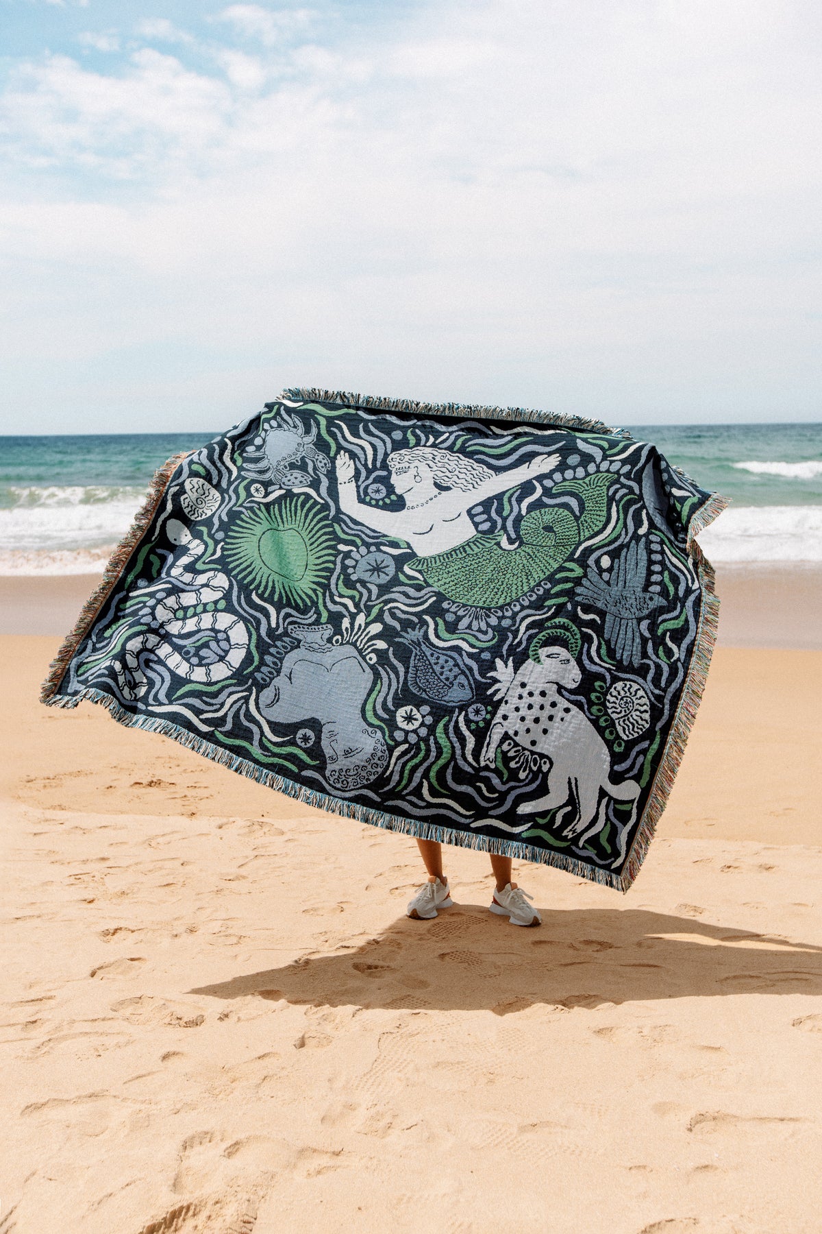 A person stands on a sandy beach holding up the Mythos Throw in Navy, featuring abstract designs inspired by Greek mythology. The ocean and sky are visible in the background.