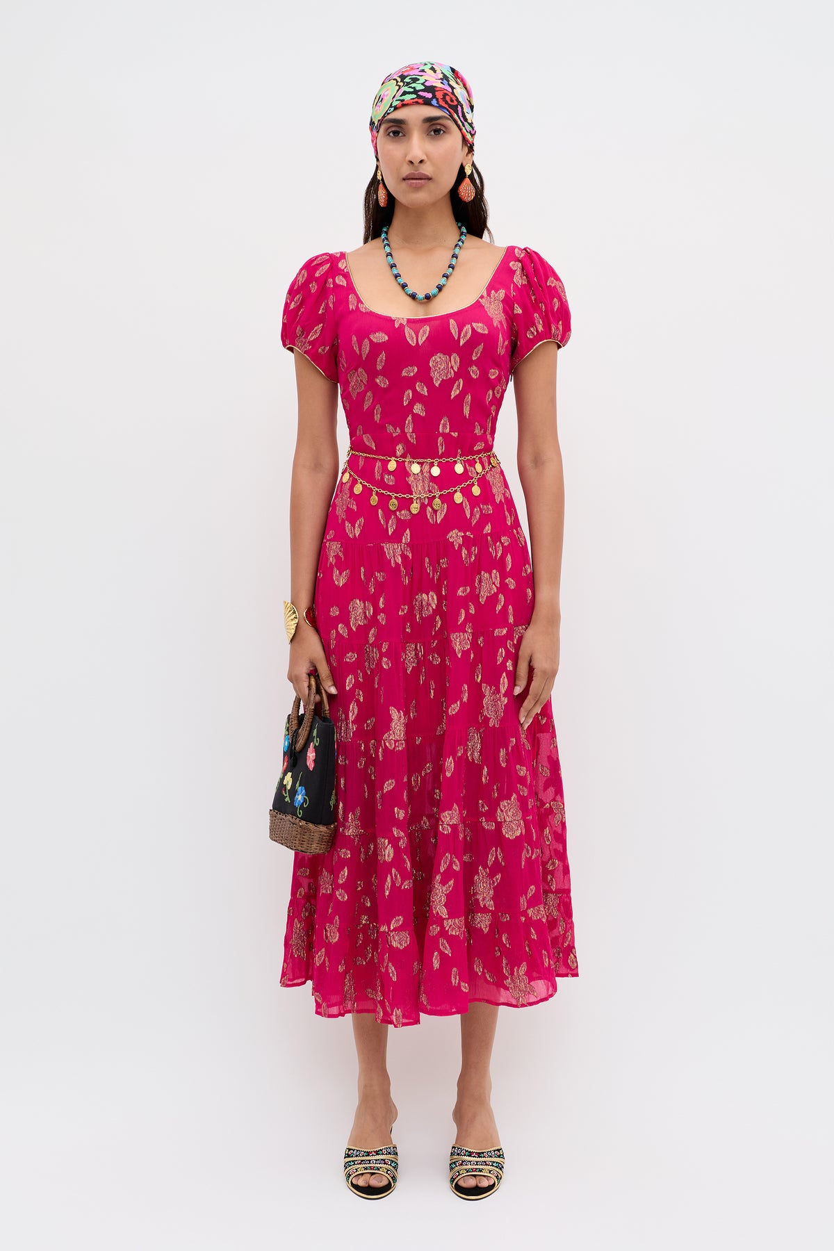 A woman wears the Remi - Floral Spark Jacquard Magenta Pink midi dress with a colorful headscarf, beaded necklace, gold belt, black handbag, and embellished sandals against a white background.
