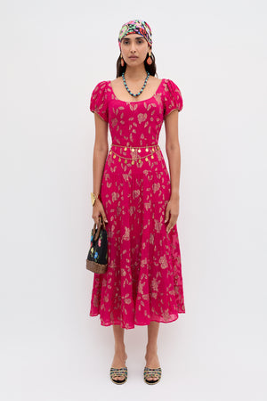 A woman wears the Remi - Floral Spark Jacquard Magenta Pink midi dress with a colorful headscarf, beaded necklace, gold belt, black handbag, and embellished sandals against a white background.