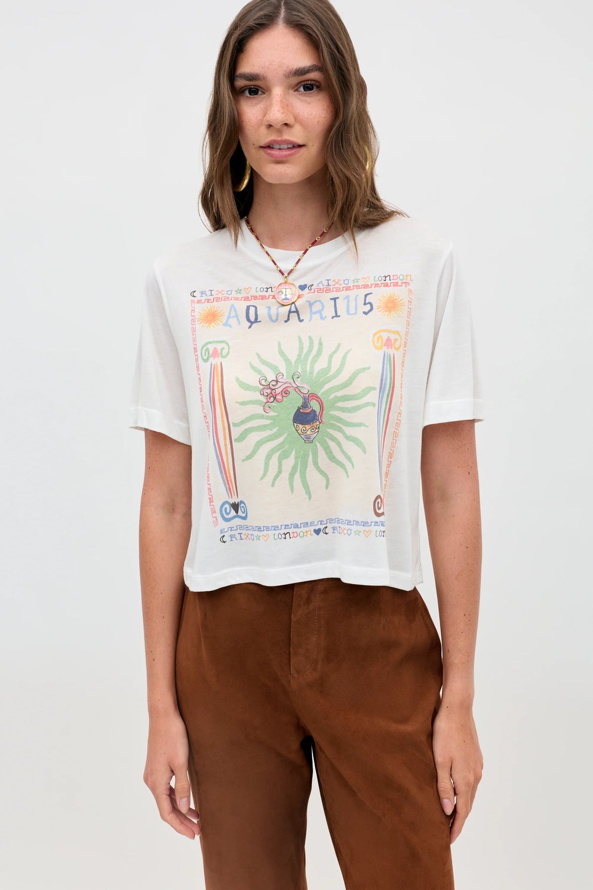 A woman with straight brown hair wears the "Ria - Aquarius" white T-shirt and cropped brown pants, standing against a plain light background.