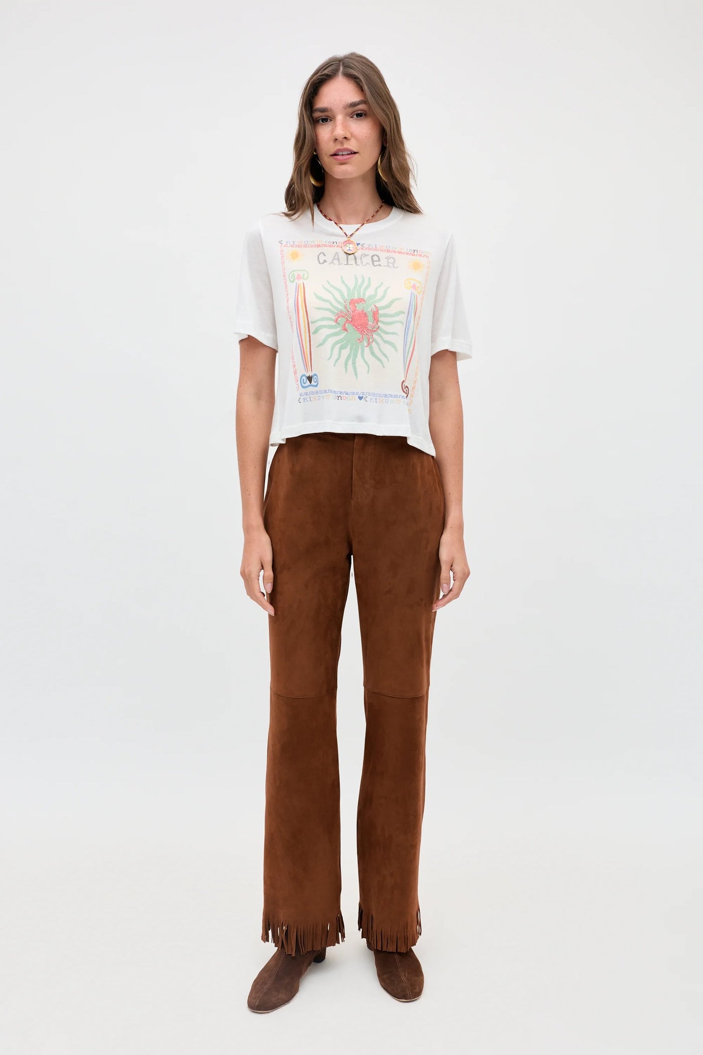 A woman stands facing forward against a plain white background, wearing the Ria - Cancer white graphic t-shirt with a subtle Cancer illustration, paired with high-waisted brown suede pants featuring a cropped hem and fringe, and brown shoes.