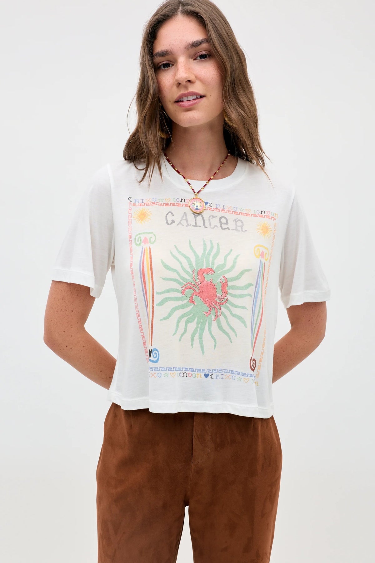 A woman with long brown hair stands against a plain light background, wearing the Ria - Cancer t-shirt. The white top features a vibrant Cancer zodiac sign graphic with a bold red crab illustration and a cropped hem, paired with brown pants.