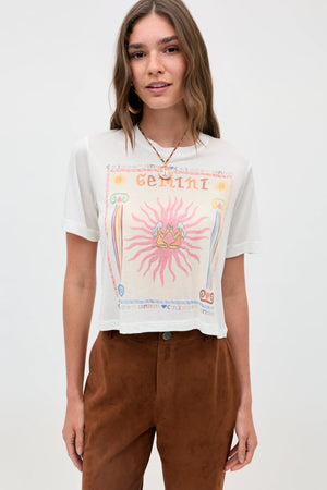 A woman stands facing the camera against a plain light background, wearing the "Ria - Gemini" white sheer jersey t-shirt featuring a colorful Gemini illustration and paired with brown pants.