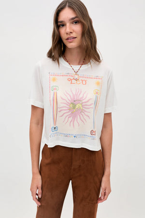 A woman with shoulder-length brown hair wears the Ria - Leo T-shirt, which features a vibrant, mystical Leo design, paired with brown pants. She stands against a plain light background, looking at the camera with a slight smile.