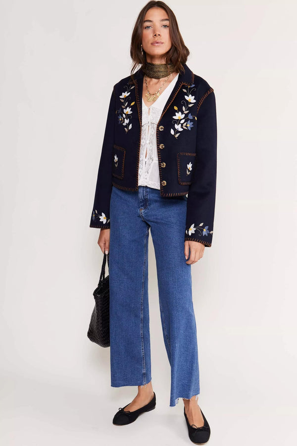 A woman stands against a plain background wearing a navy embroidered jacket, white blouse, wide-leg jeans, black flats, and layered necklaces featuring the Channing - Gold. She accessorizes with a vintage scarf and holds a black textured handbag.