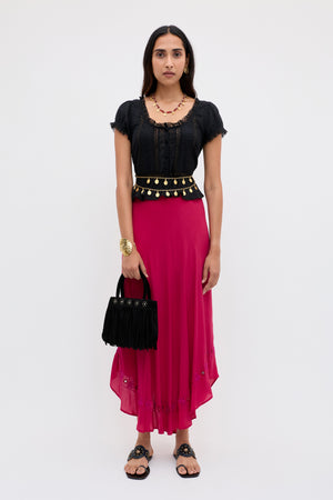 A woman stands facing forward against a plain light background, wearing the Rudy - Mirror Embellishment Magenta Pink skirt. She pairs it with a black top, black sandals, jewelry, and holds a black fringed handbag.