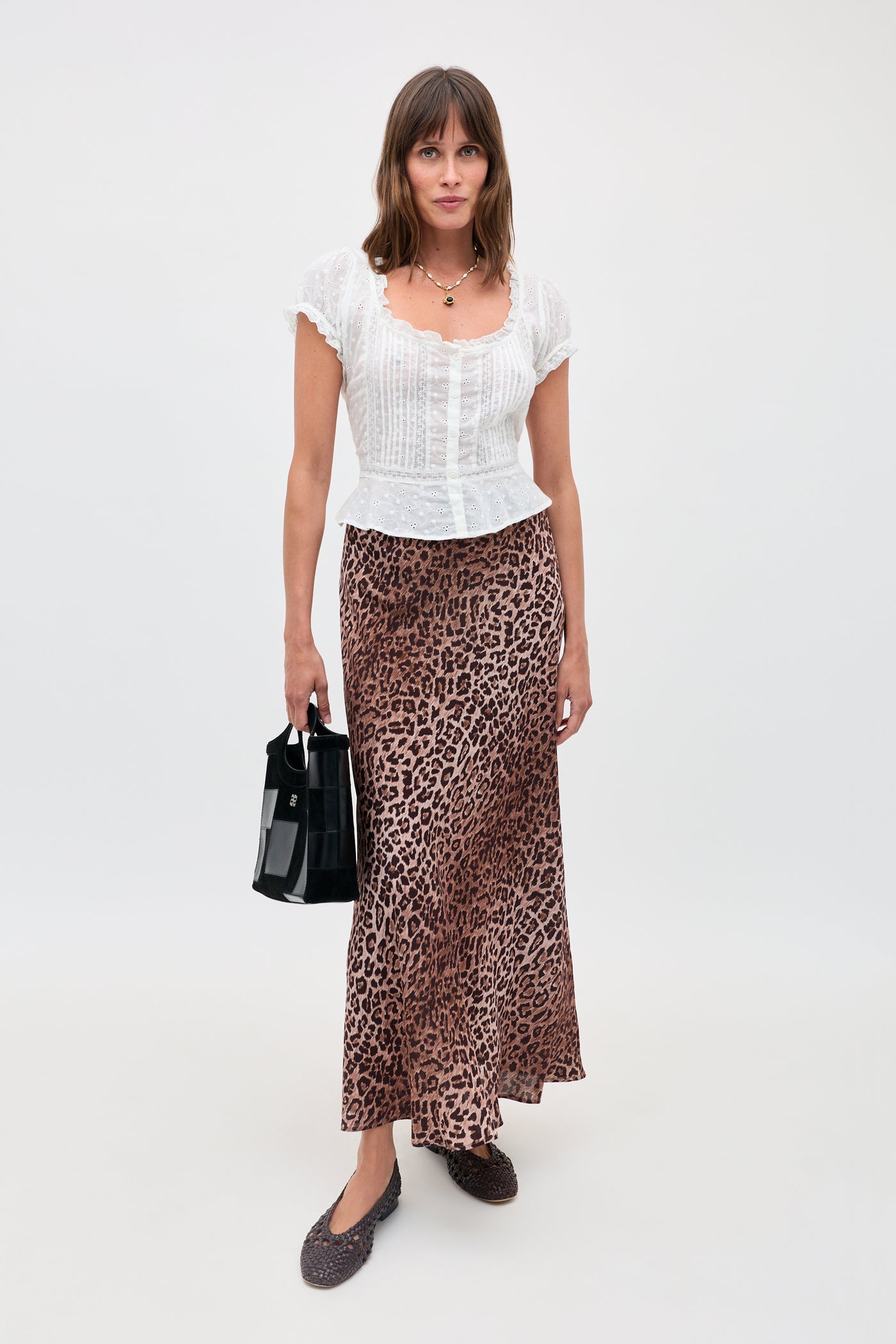 A woman stands against a plain background wearing the Sandi - Ivory short-sleeve blouse with delicate lace inserts, a long leopard-print skirt, black flats, and holding a black handbag for effortless everyday style.