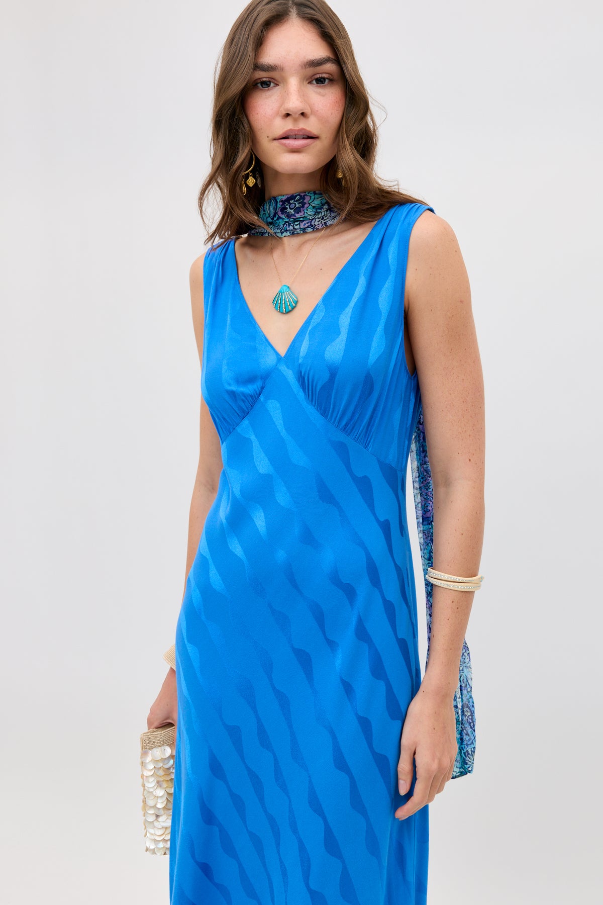 A woman with wavy brown hair wears the sleeveless, V-neck Sandrine - Wave Jacquard Blue dress styled with a patterned scarf, white bangles, a shell necklace, and a beaded clutch.