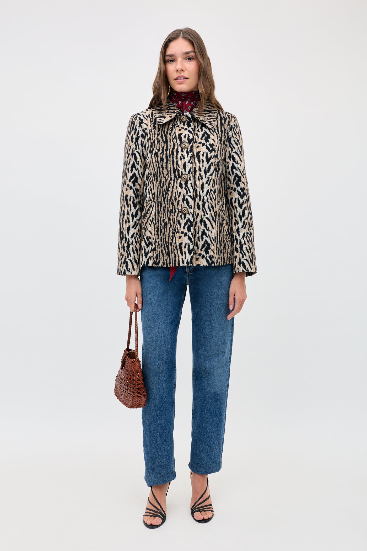 A woman stands facing forward against a plain background, wearing the Tommie - Bohemia Leopard jacket, paired with blue jeans, black strappy heels, and holding a brown woven handbag. She has straight brown hair and a neutral expression.