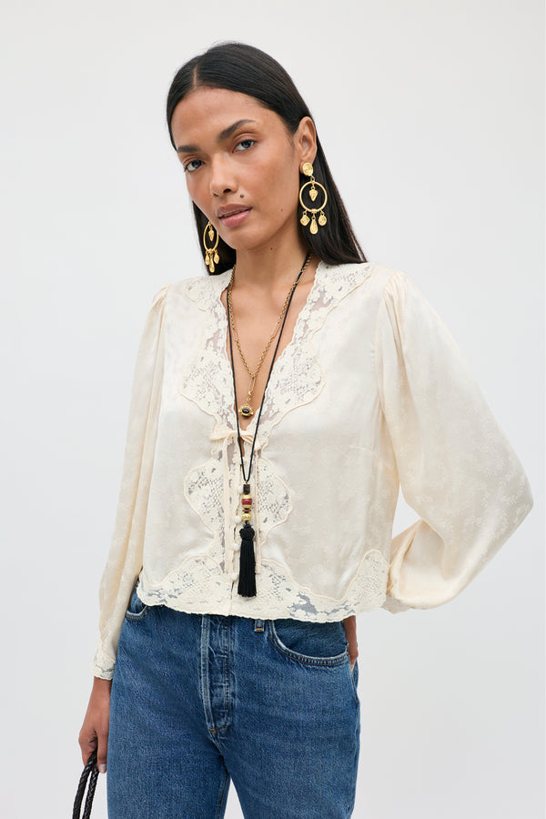 A woman wearing the Tove - Floral Trail Jacquard Champagne blouse with long sleeves, styled with blue jeans, layered necklaces, and large gold earrings, stands against a plain light background.