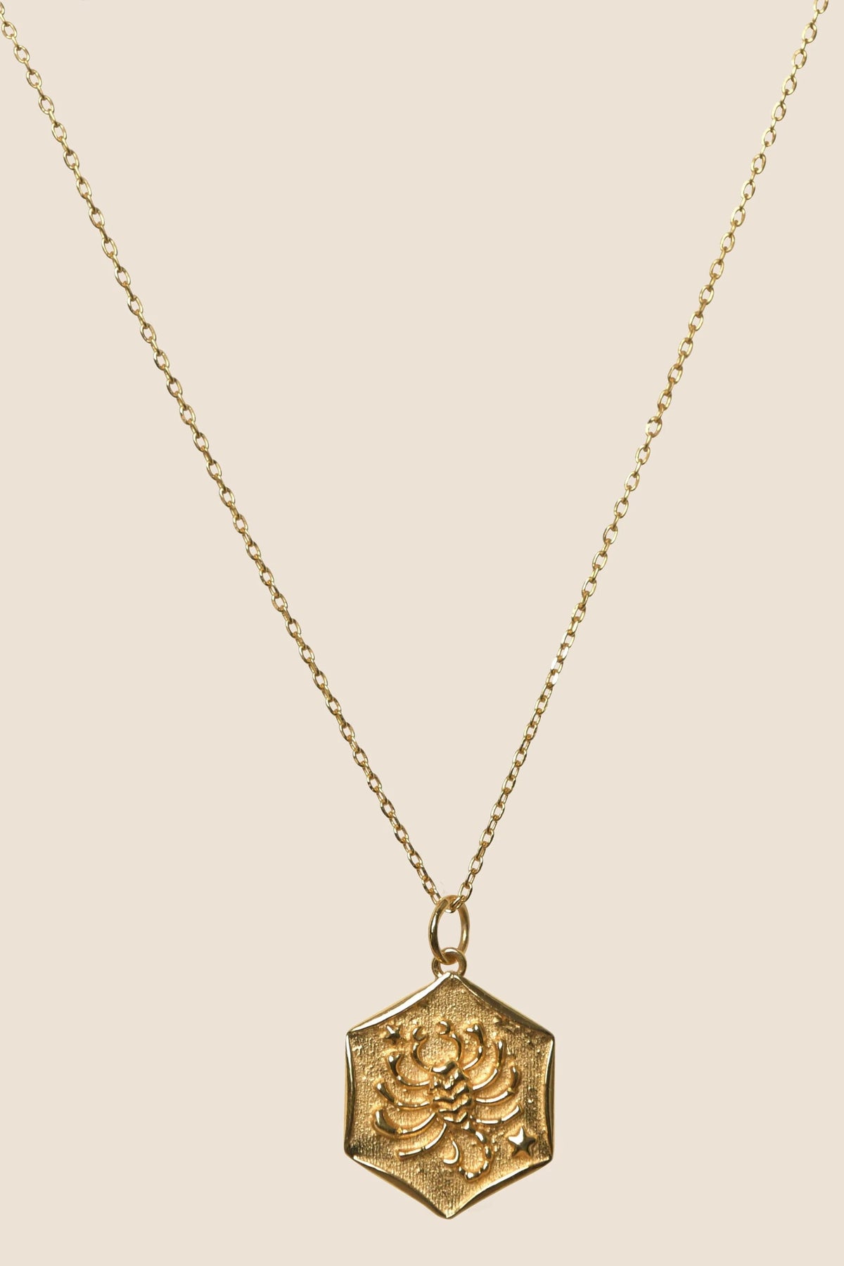 A delicate gold vermeil chain necklace, Zodiac - Scorpio, with a hexagonal pendant featuring an embossed floral design, displayed against a plain beige background—a subtle nod to zodiac elegance.