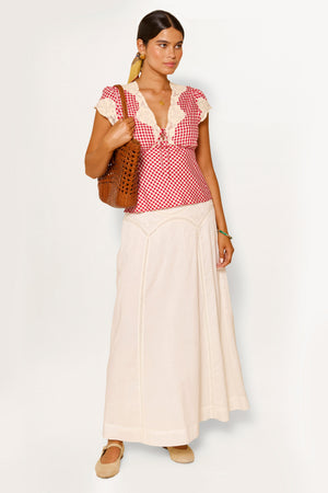 A woman stands against a white background wearing the Vienne - Pansy Embroidery Ivory skirt, paired with a red and white checkered blouse, beige shoes, gold earrings, and holding a brown woven bag.