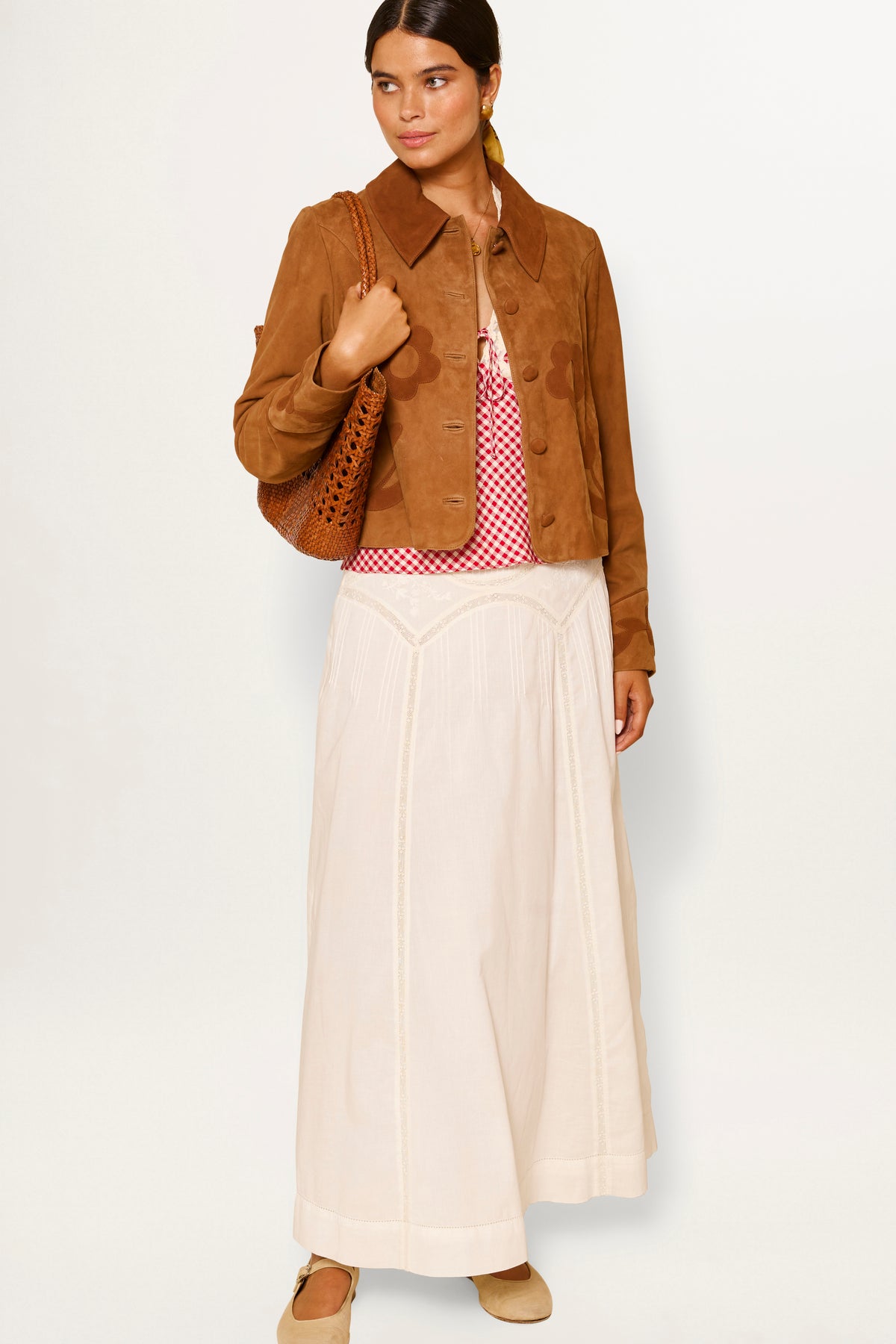 Woman wearing the Vienne - Pansy Embroidery Ivory vintage-inspired white skirt with a brown suede jacket, red and white patterned top, beige shoes, and holding a brown woven bag against a plain white background.