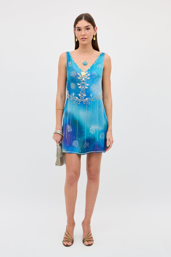 A woman wearing the Kaylor - Jessica Alexandra Blue, a sleeveless mini dress featuring blue ombré hues with silver embellishments and delicate Aari embroidery, accessorized with a small silver bag, gold sandals, and gold jewelry, stands against a plain white background.